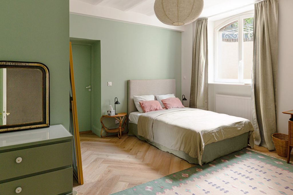 Chambre aux murs vert sauge avec lit double, coussins rose poudré et sol effet bois à bâtons rompus.