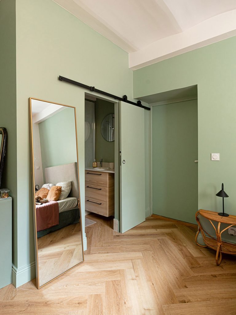 Chambre aux murs vert sauge avec salle d’eau attenante, porte coulissante, grand miroir et sol effet bois à bâtons rompus.