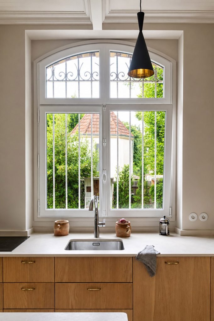 Cuisine beige et bois aux façades en chêne et poignées laiton, avec évier installé sous une grande fenêtre cintrée.