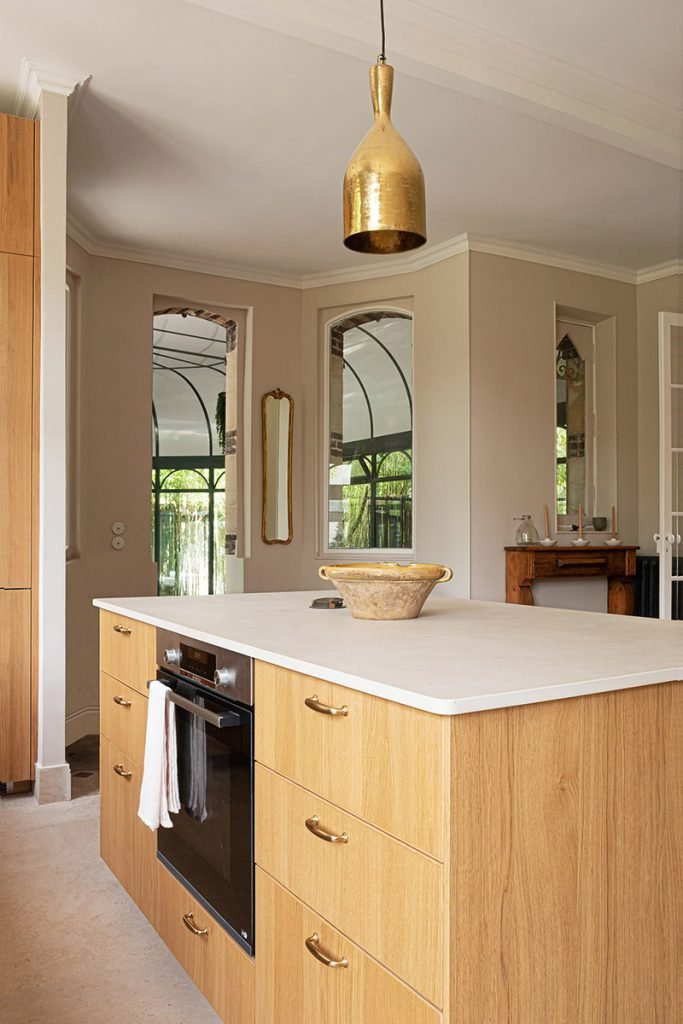 Îlot central en chêne avec plan de travail en Dekton dans une cuisine beige et bois, avec four intégré, plan de travail clair et suspension en laiton.