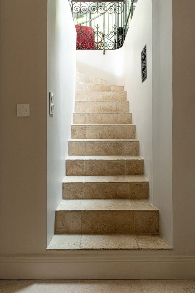 Escalier en travertin vu depuis le bas, avec murs beiges et garde-corps ancien en fer forgé.