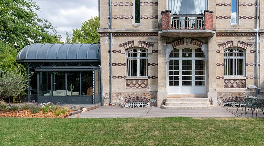 Façade en brique d’une maison ancienne avec verrière latérale, terrasse et jardin.