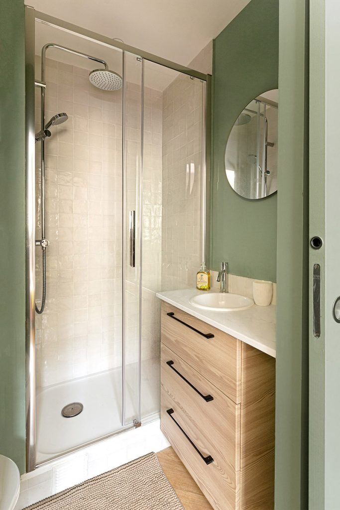 Salle d’eau vert sauge avec douche vitrée, meuble vasque en bois, carrelage beige et miroir rond.