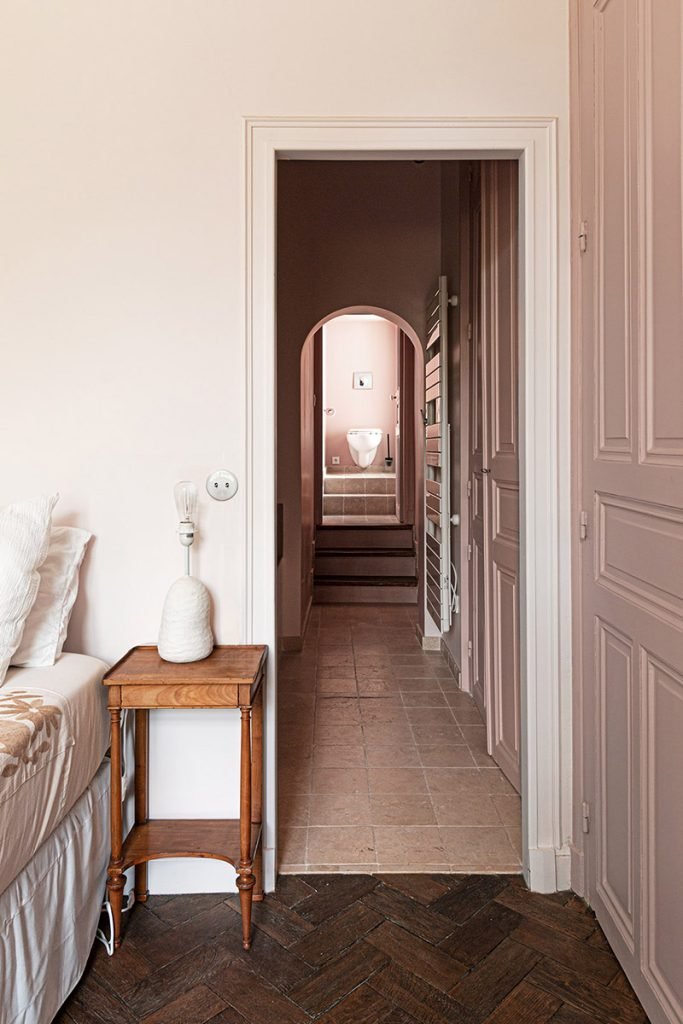 Vue en enfilade d’une suite parentale rose poudré vers la salle de bains, avec parquet à bâtons rompus et sol en travertin conservés.