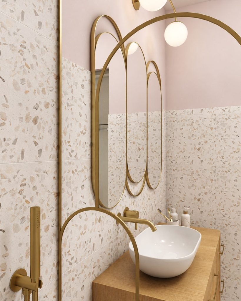 Salle d’eau féminine en terrazzo clair et rose quartz, avec meuble vasque en bois, miroirs arrondis et robinetterie en laiton.