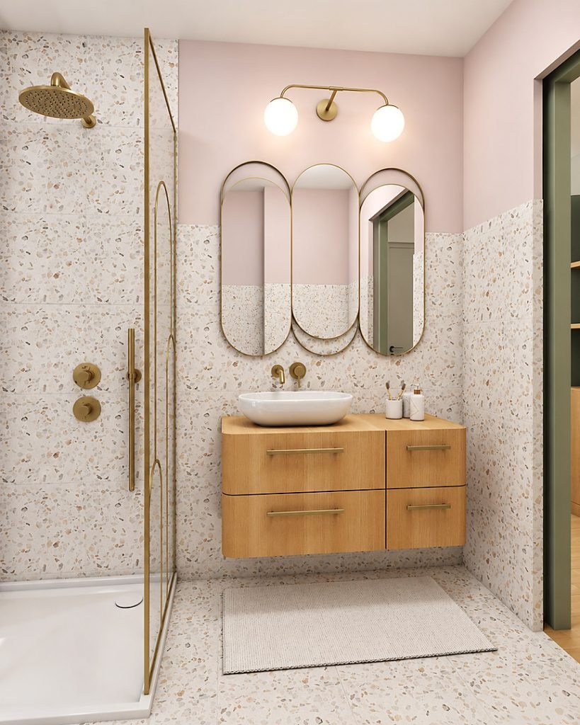 Vue d’ensemble de la salle d’eau rose et terrazzo, avec douche, paroi en laiton, meuble vasque suspendu en bois et miroirs arrondis.